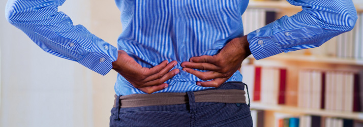 A person in a blue striped shirt holding their lower back in pain while standing near a bookshelf indoors.