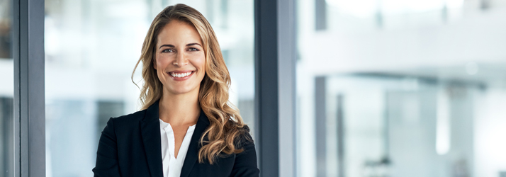 A smiling, professional female Medway chiropractor wearing a dark blazer, standing in a bright, modern office setting.