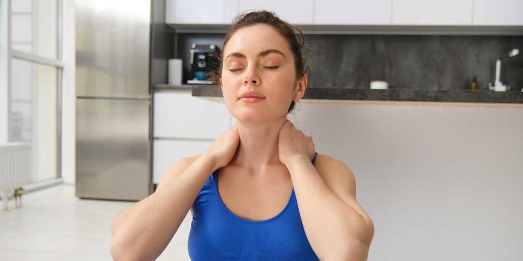 Woman in a blue tank top massaging her neck with eyes closed during a home neck pain recovery exercise routine.