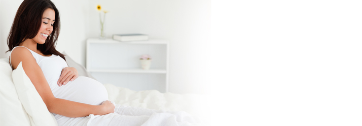 Smiling pregnant woman resting comfortably on a bed, holding her belly while considering safe chiropractic care in Medway area.