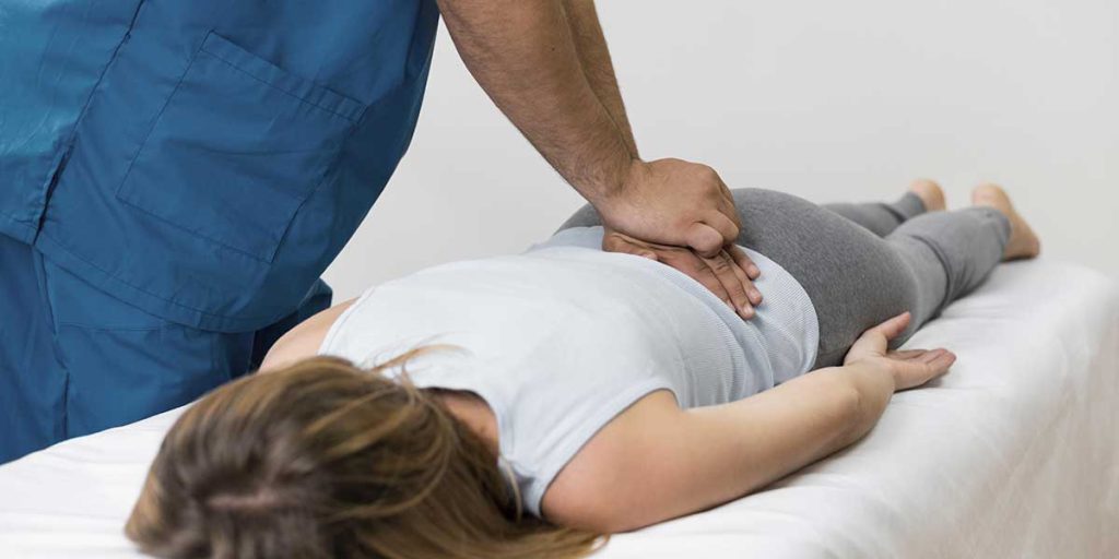 Healthcare practitioner applying targeted manual pressure to a patient's lower back while lying face down on a treatment table.